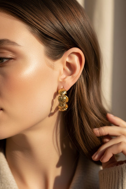 Gold hoop earrings featuring a repeating heart-shaped link design, worn by a woman with side-swept hair.