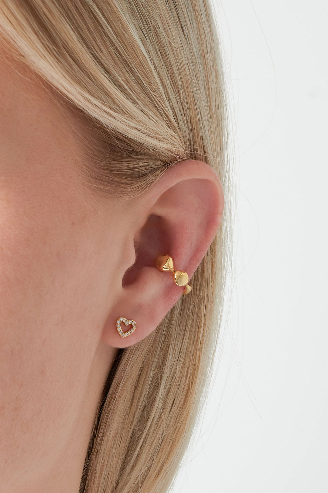 Model wearing gold wavy ear cuff on upper ear and heart-shaped diamond stud earring on earlobe