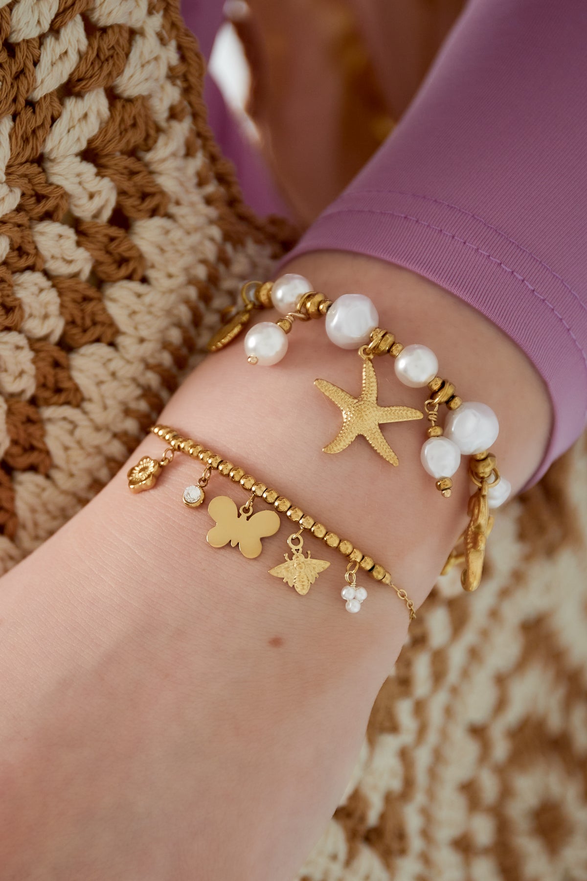 Wrist adorned with layered bracelets including a gold-tone charm bracelet with butterfly, bee, flower, crystal, and pearl charms, combined with a pearl beaded and starfish charm bracelet