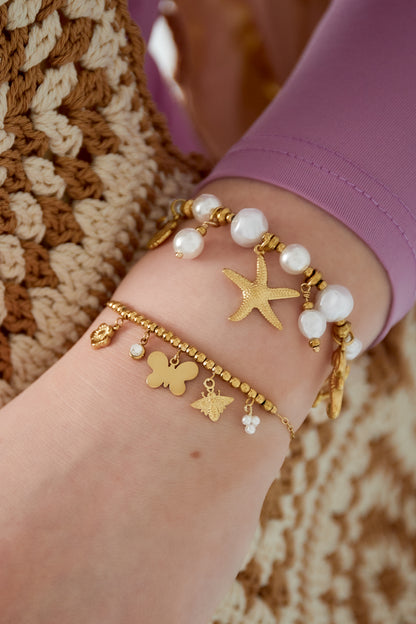 Wrist adorned with layered bracelets including a gold-tone charm bracelet with butterfly, bee, flower, crystal, and pearl charms, combined with a pearl beaded and starfish charm bracelet