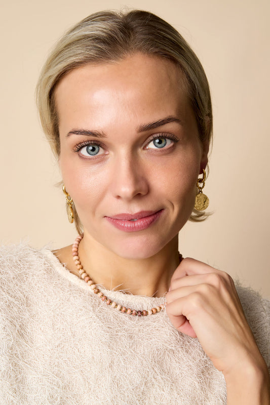 Woman wearing a pink stone beaded necklace and gold coin drop earrings against beige background