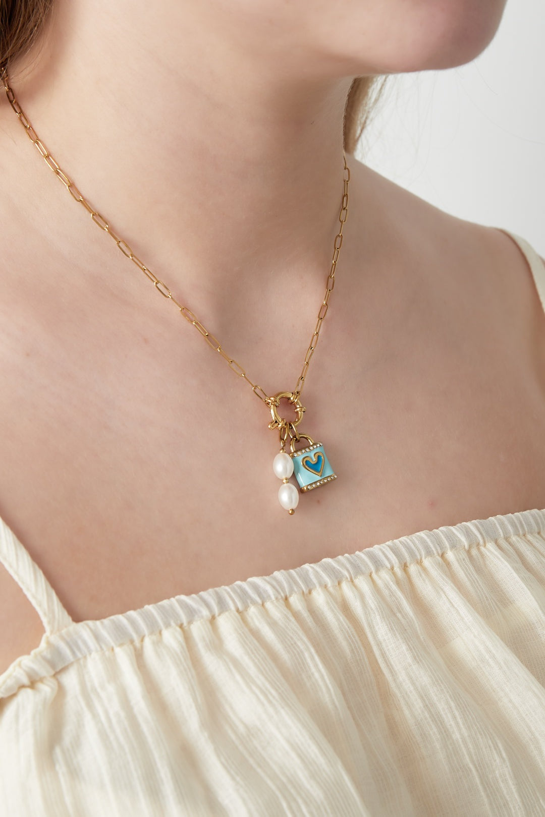 Close-up of the gold paperclip chain necklace with blue heart pendant and pearl charm worn by a woman in an off-white top