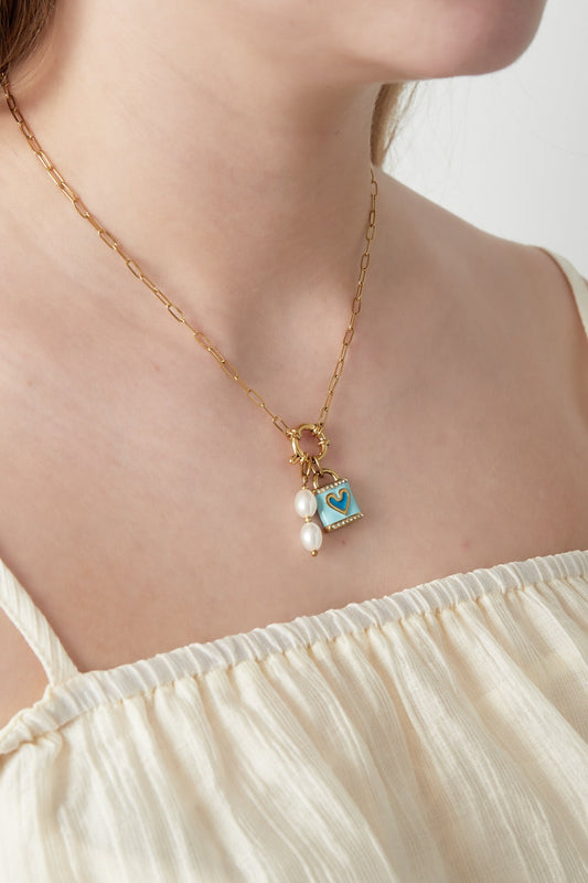 Close-up of the gold paperclip chain necklace with blue heart pendant and pearl charm worn by a woman in an off-white top