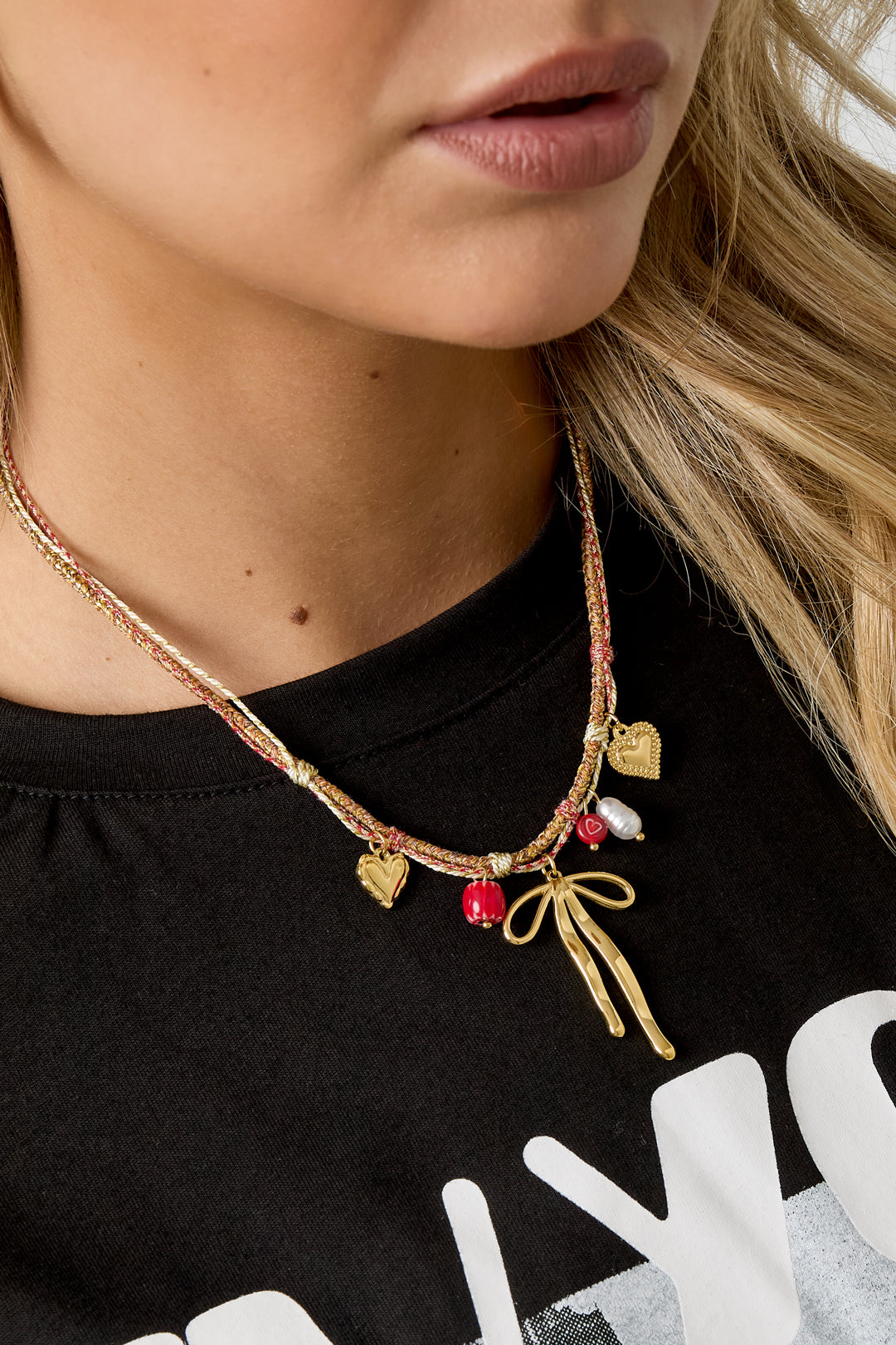Model wearing a playful gold-tone bow knot charm necklace with hearts and beads over a black shirt with white print