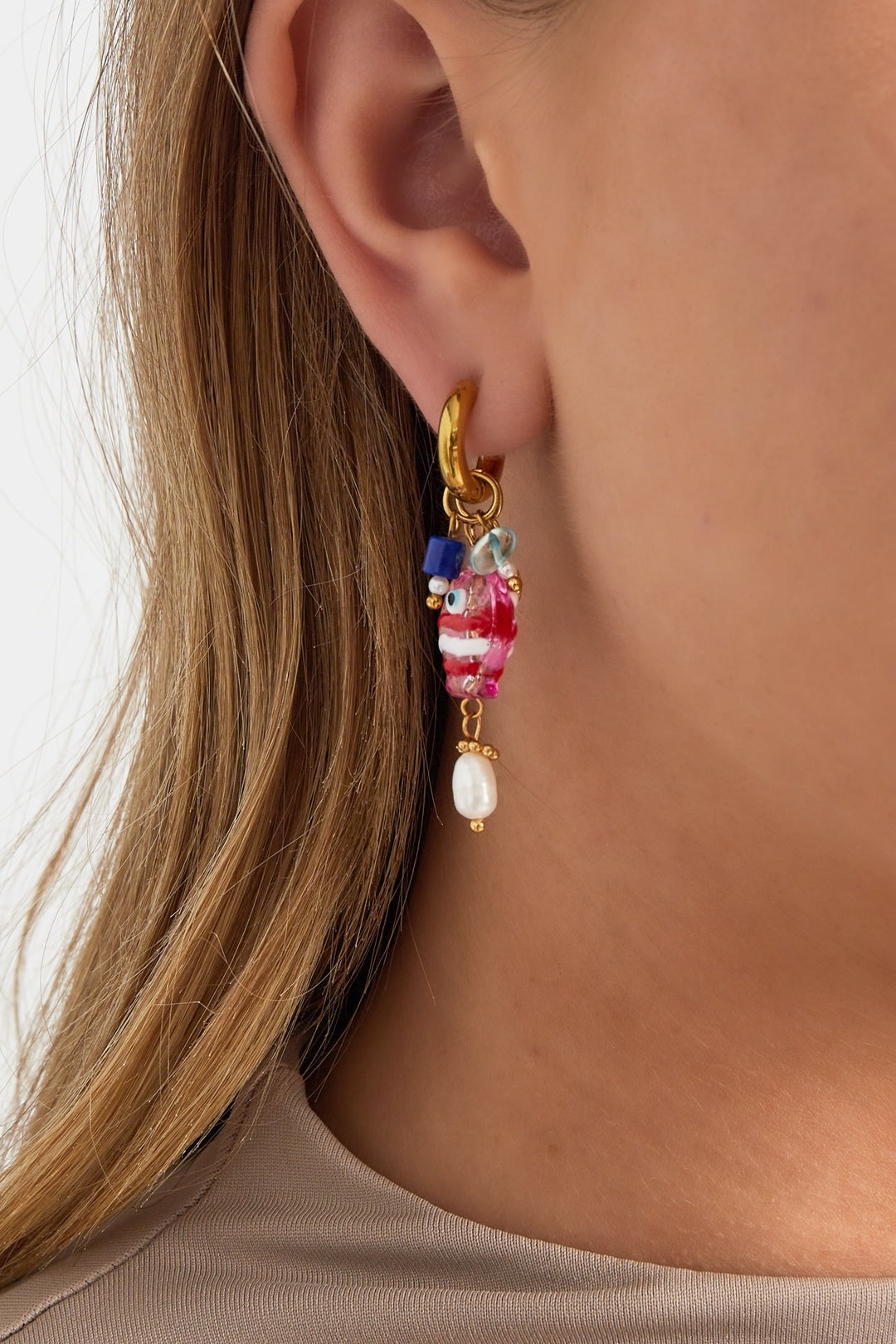 Close-up of a woman's ear wearing a gold-tone hoop earring adorned with a pink and white striped resin fish charm, blue beads, and a faux pearl drop