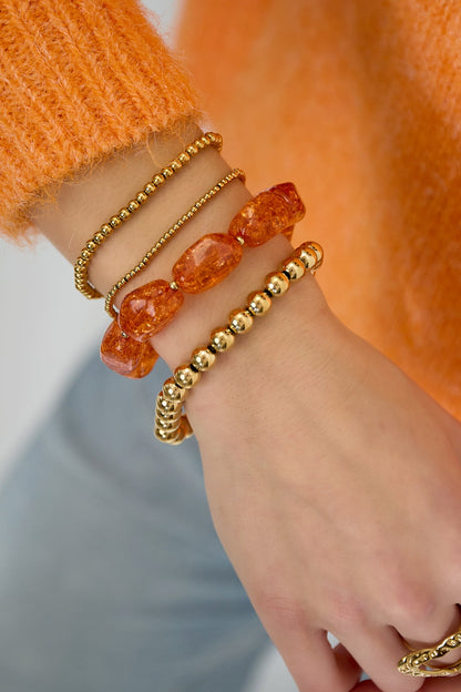 Model wearing layered bracelets featuring large orange beads and small gold beads with orange sweater and blue jeans