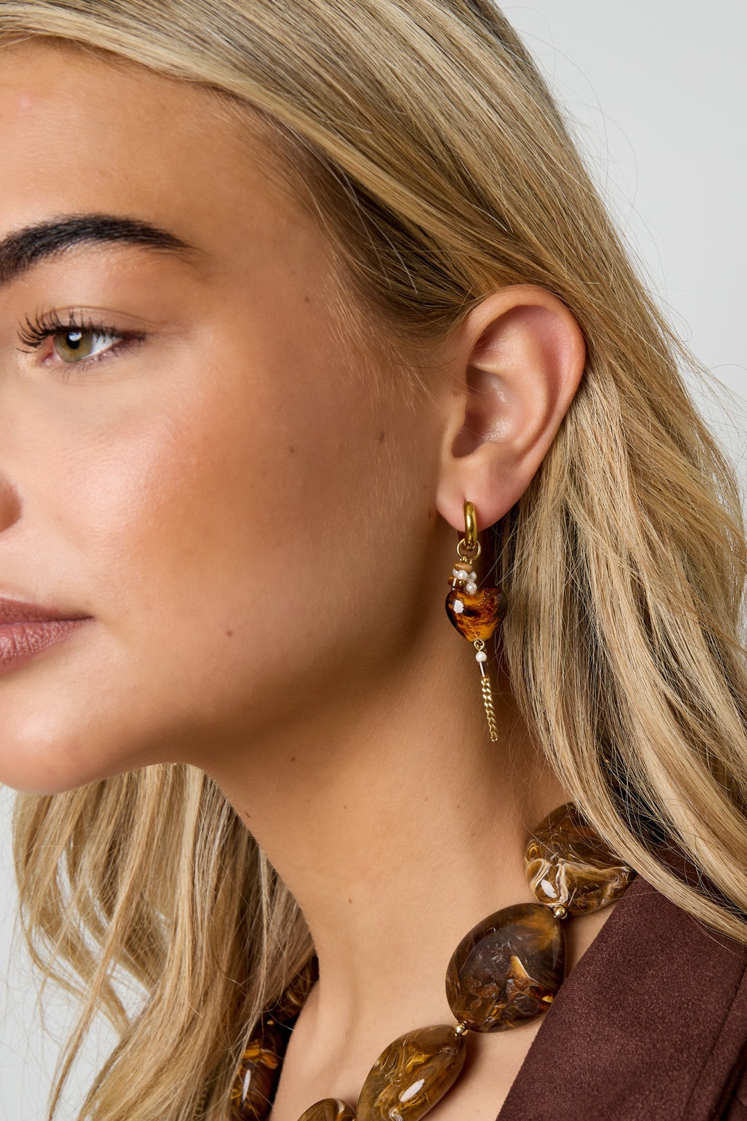 Close-up of a woman wearing a gold hoop earring with an amber tortoiseshell heart charm, pearl accents, and a dangling chain, styled with matching necklace