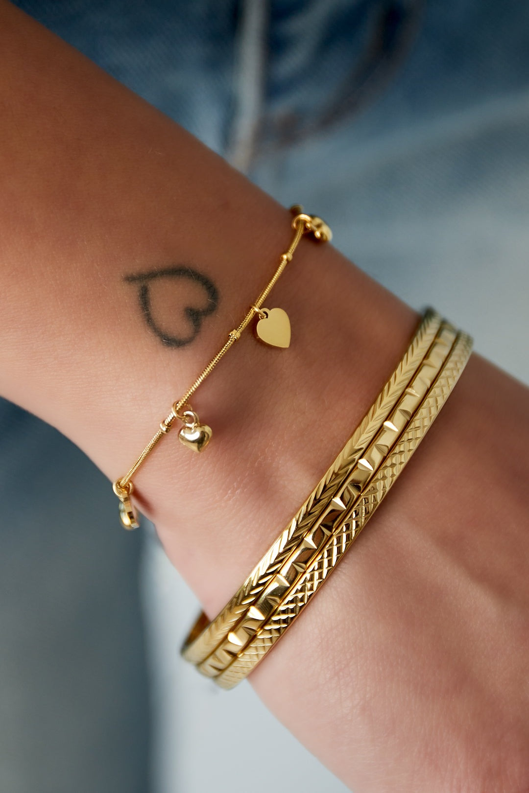 Close-up of a wrist wearing the gold-tone heart charm bracelet paired with textured gold bangles, visible small heart tattoo on wrist