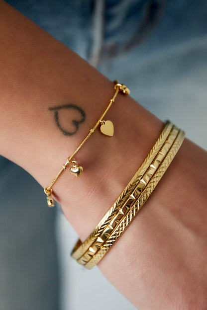 Close-up of a wrist wearing the gold-tone heart charm bracelet paired with textured gold bangles, visible small heart tattoo on wrist