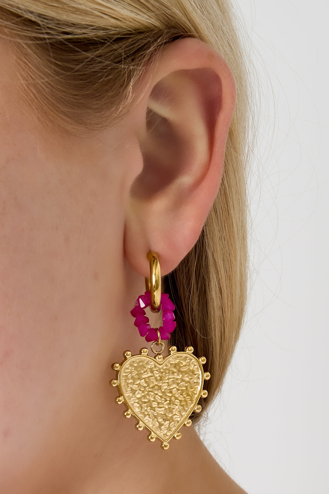 Close-up of a woman's ear wearing gold hoop earrings with textured heart pendants and pink beaded connectors showcasing size and style