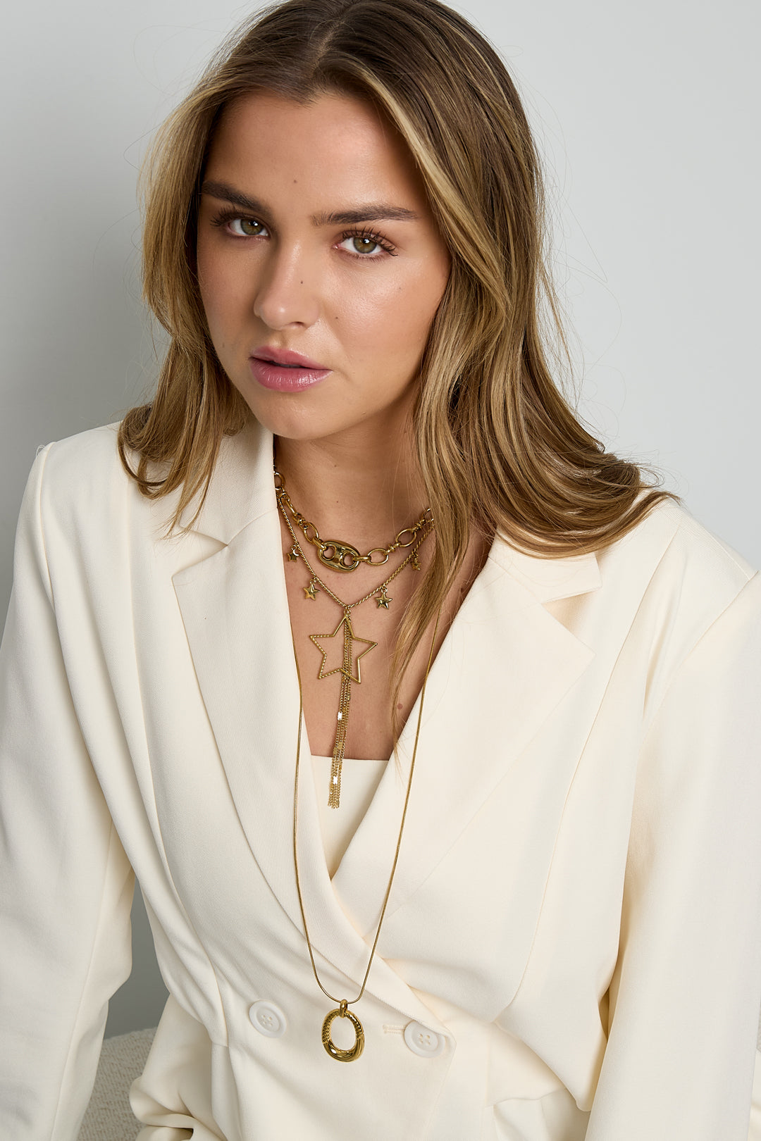 Model wearing gold star charm necklace layered with chunky chain and long gold pendant necklace with cream blazer