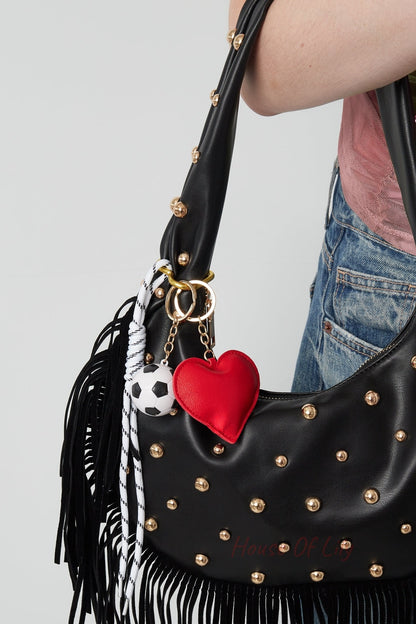 Woman carrying black studded leather bag with playful red heart and soccer ball bag charm attached on gold rings