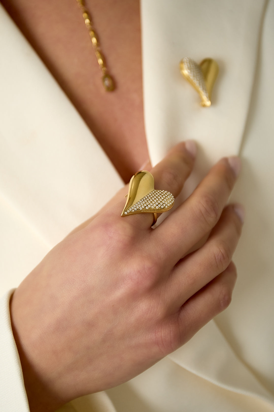 Close-up of hand wearing the gold heart-shaped rhinestone ring paired with matching heart brooch on cream blazer