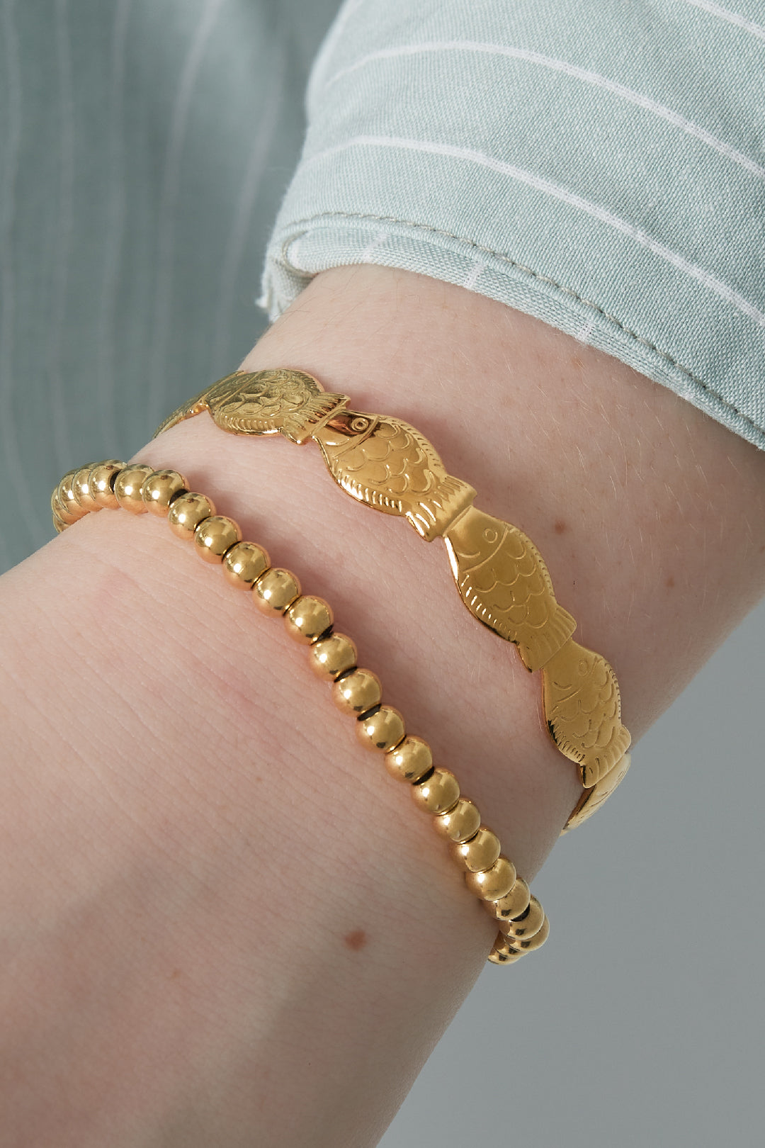 Close-up of wrist wearing gold beaded stretch bracelet paired with a gold fish-design bracelet and light blue sleeve