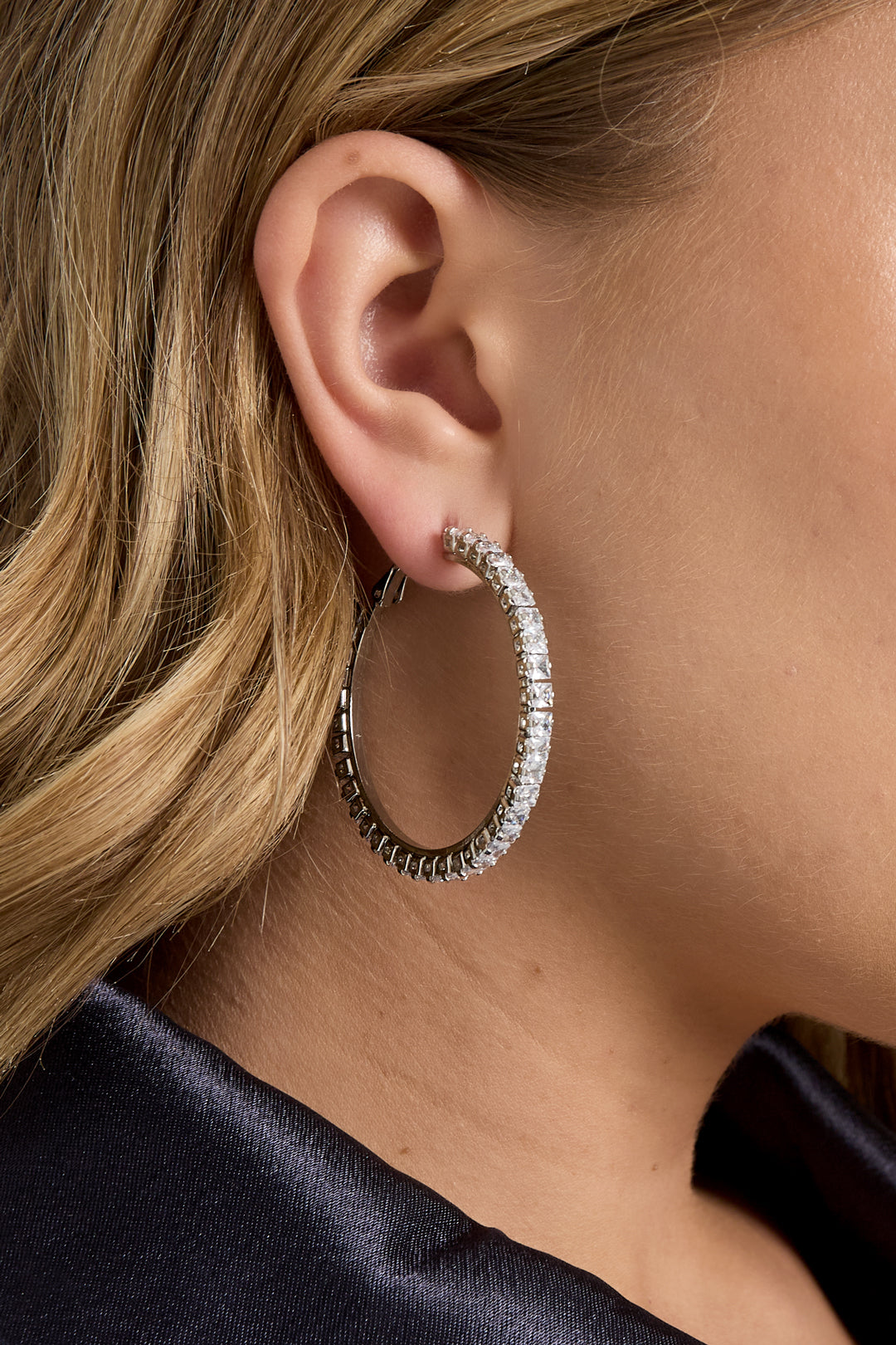 Side profile showing a woman modeling silver-tone hoop earrings with clear crystals, worn with a dark jacket.
