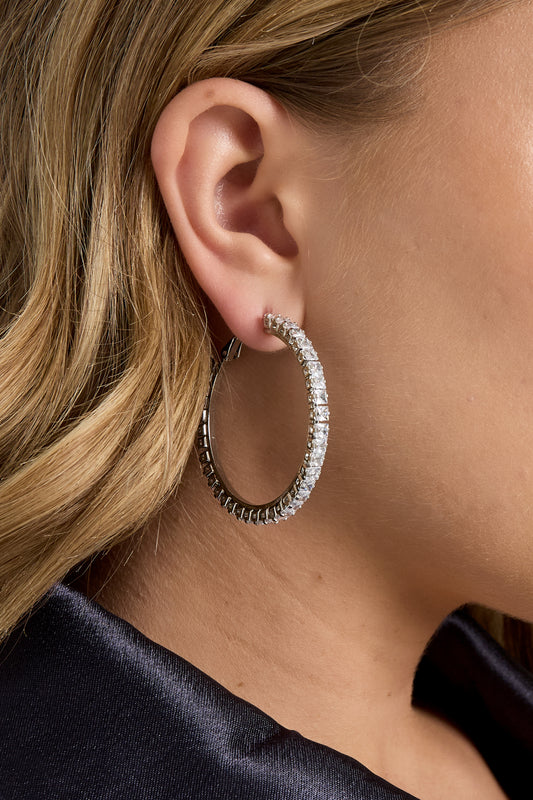 Side profile showing a woman modeling silver-tone hoop earrings with clear crystals, worn with a dark jacket.