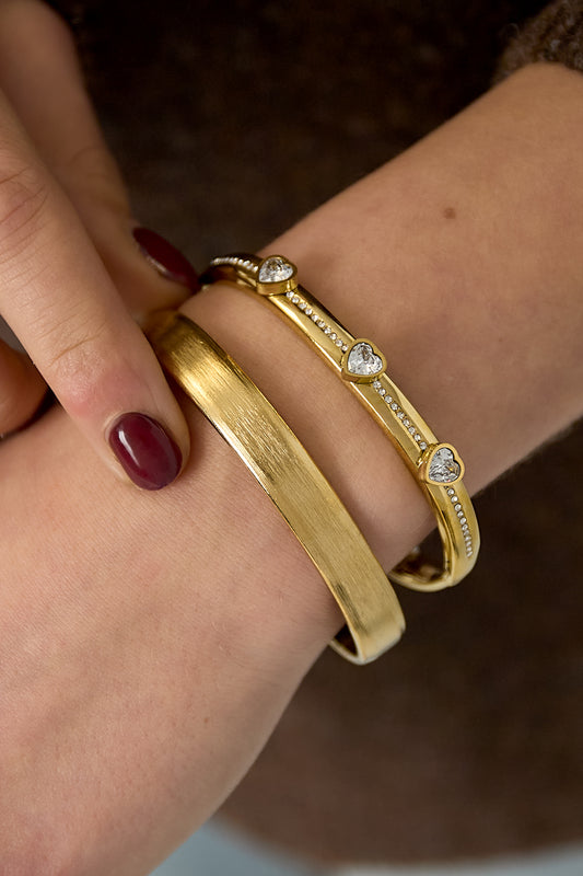 Close-up of wrist wearing gold brushed metal cuff bracelet paired with a delicate heart charm bracelet