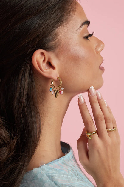 A woman wearing a gold hoop earring with colorful ceramic heart, bead, and pearl charms against a pink background.