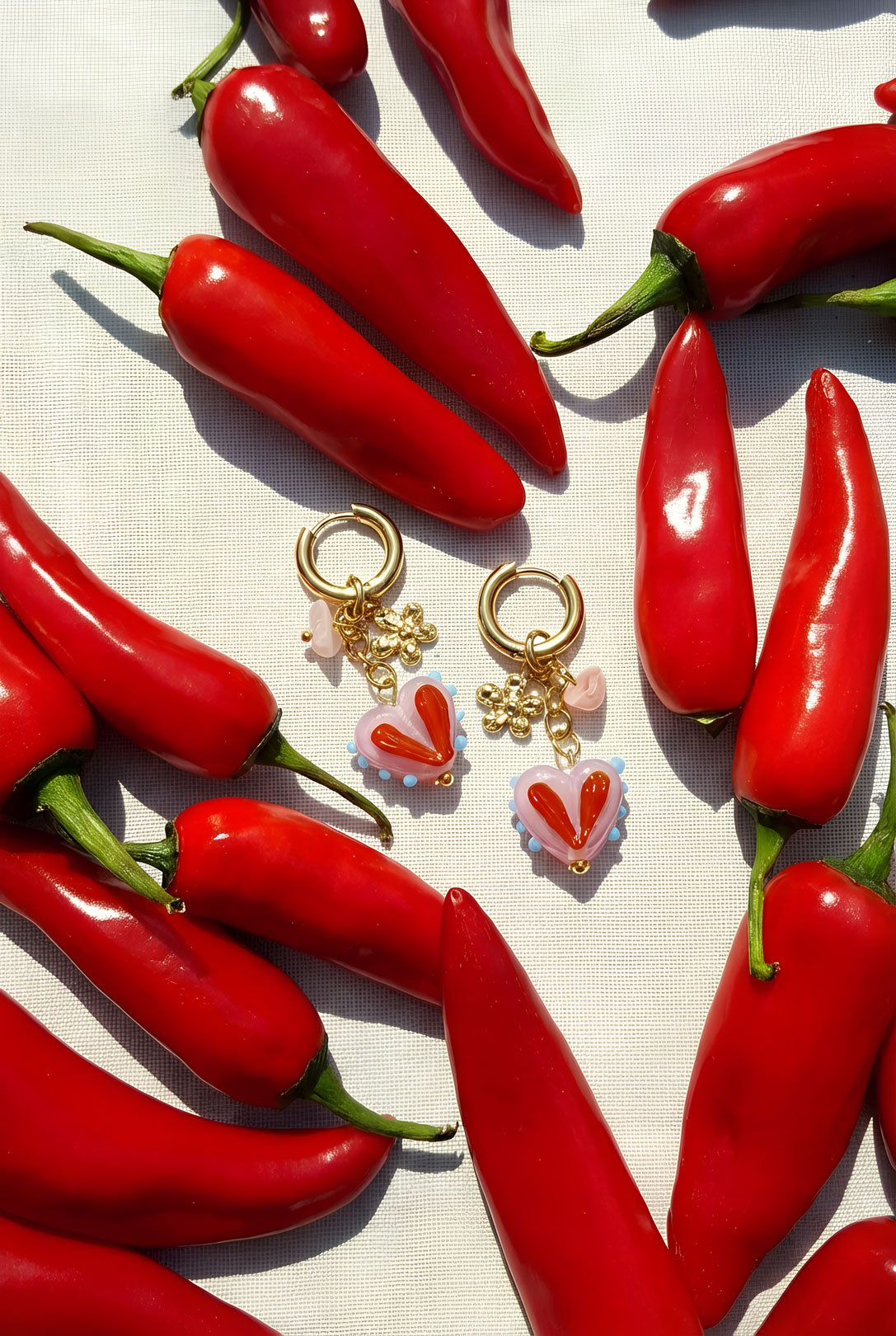 Gold hoop earrings with pink and red heart charms surrounded by fresh red chili peppers on white surface