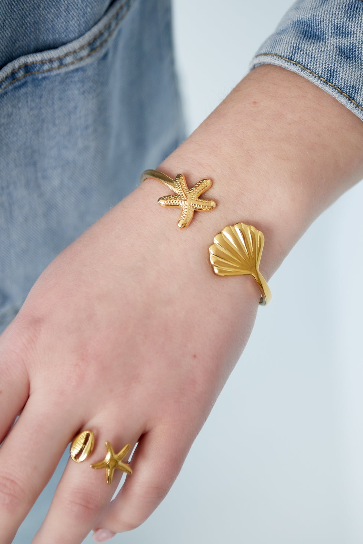 Wrist wearing gold cuff bracelet featuring starfish and seashell charms, paired with matching ring on fingers, denim jacket visible