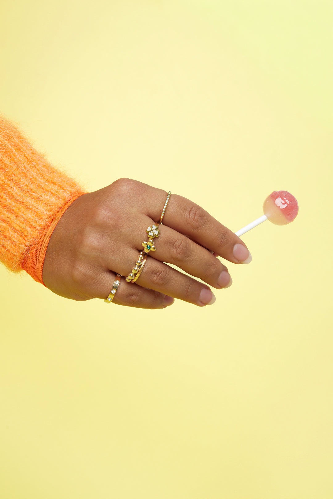 Woman's hand with multiple gold rings holding a pink and white lollipop against yellow background