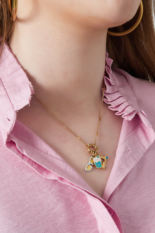 Close-up of a woman wearing a gold charm necklace featuring bubble tea, sunglasses, and scooter charms paired with a pink ruffled blouse
