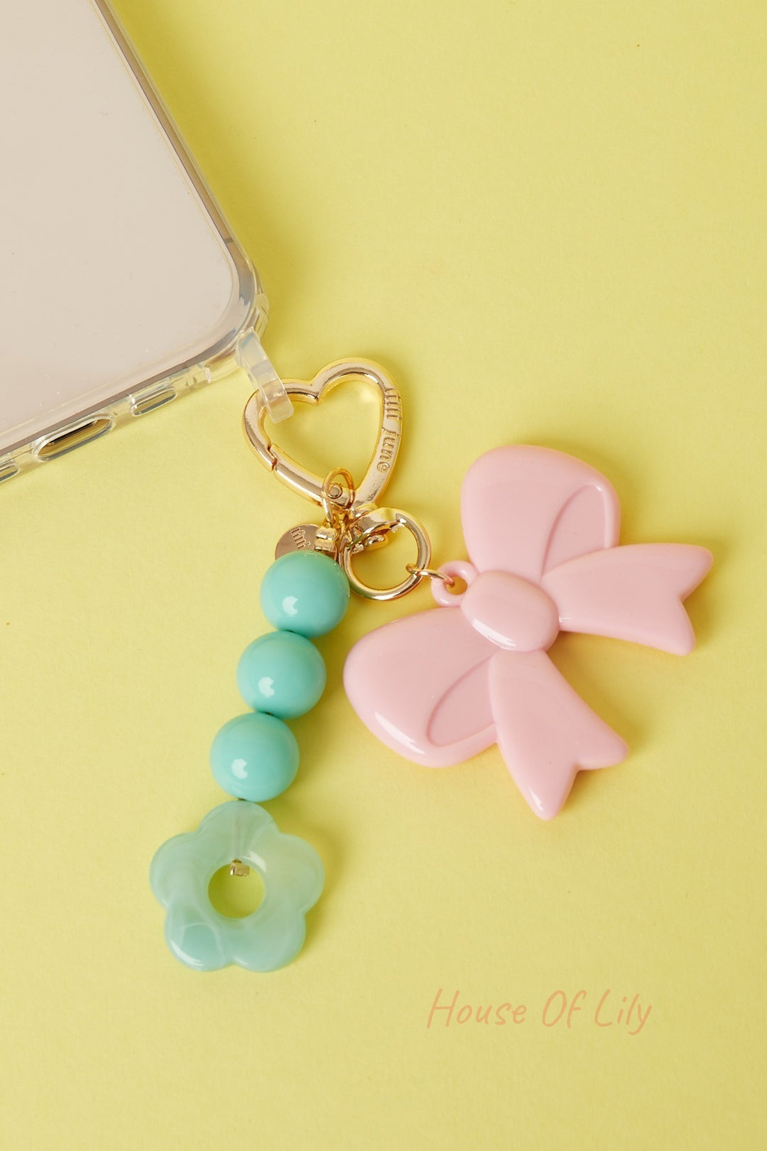Mint green acrylic flower bag charm with three round beads and pink bow charm attached to a heart-shaped gold clasp near a phone on yellow background