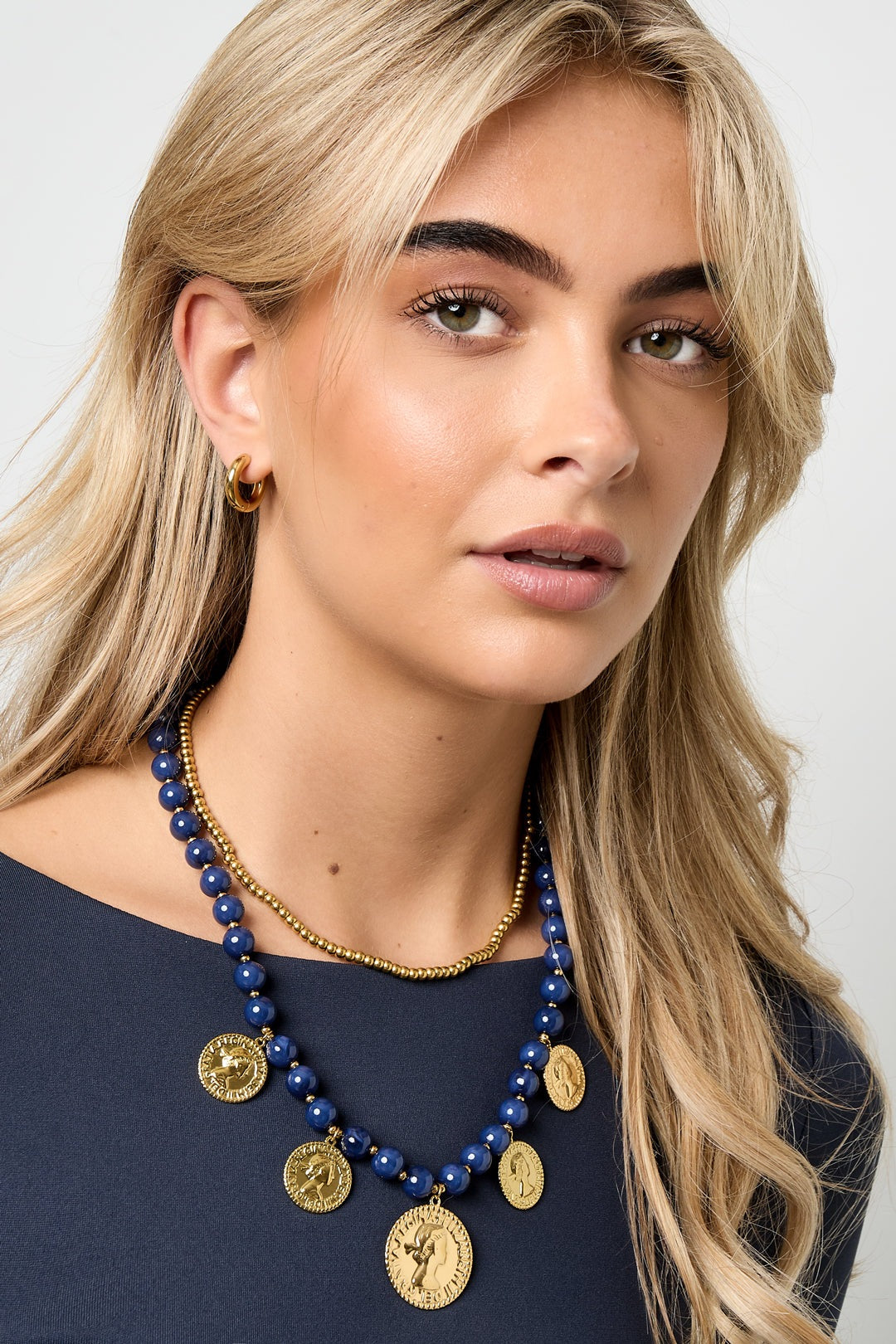 Blonde woman wearing a navy blue acrylic beaded necklace with gold-tone coin pendants paired with a gold chain choker