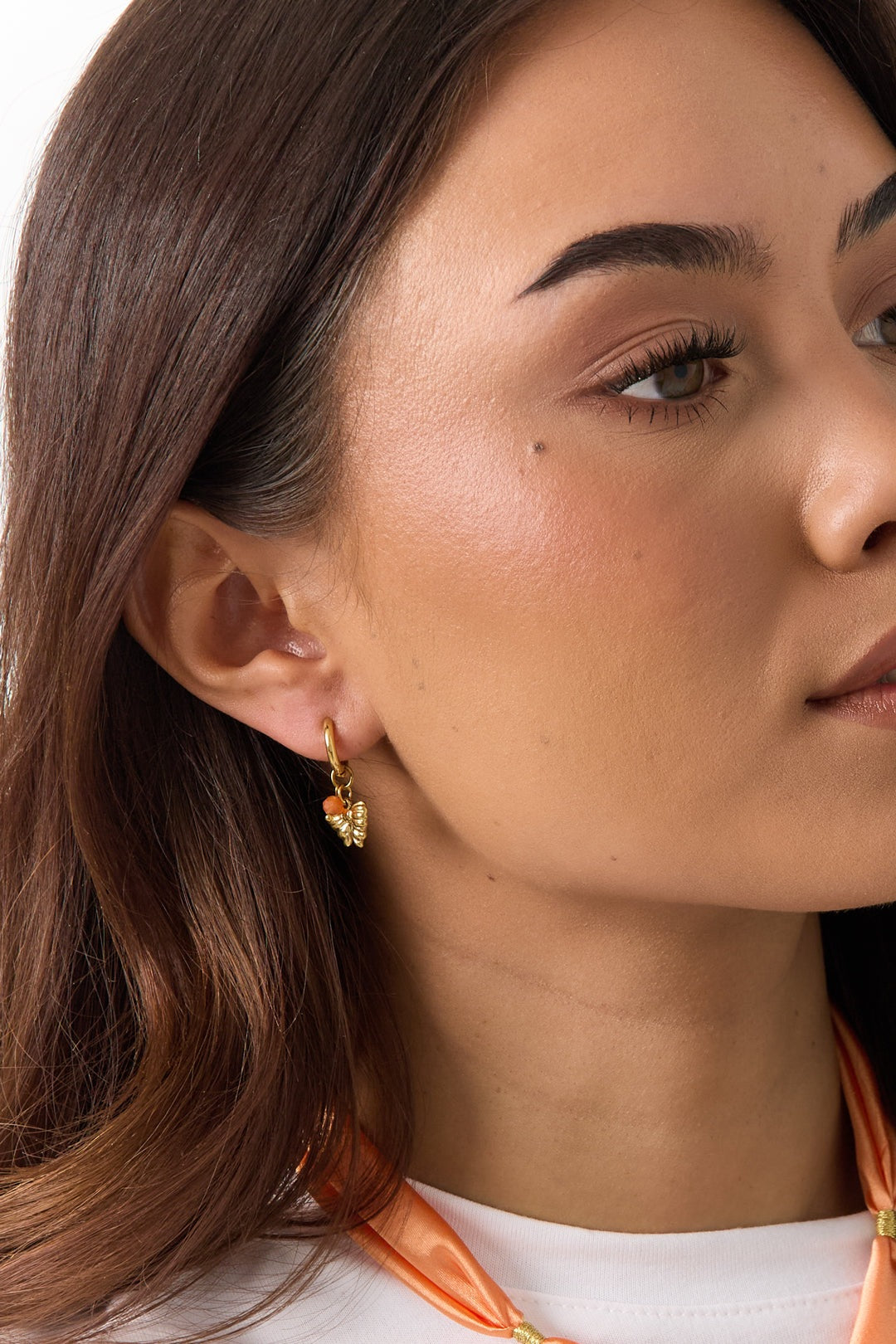 Model wearing gold hoop earrings with a small, textured golden butterfly charm and a tiny orange accent bead.