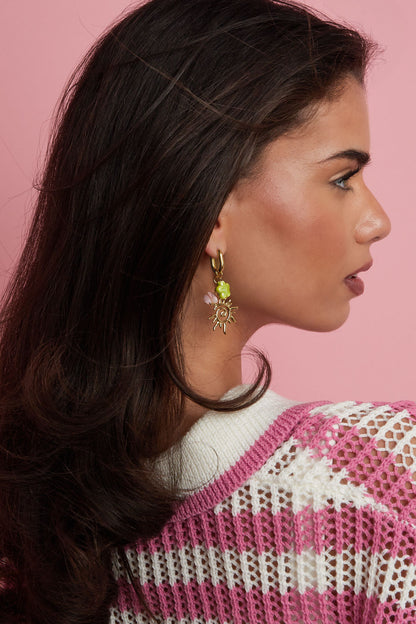 Model wearing gold-tone drop earrings featuring a sun charm, green four-leaf clover, and pink beads against a pink background
