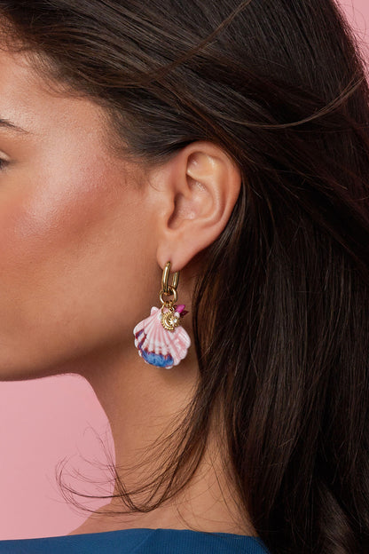 Close-up side view of woman wearing gold hoop earrings with detailed pink and blue hand-painted seashell charms against a pink backdrop