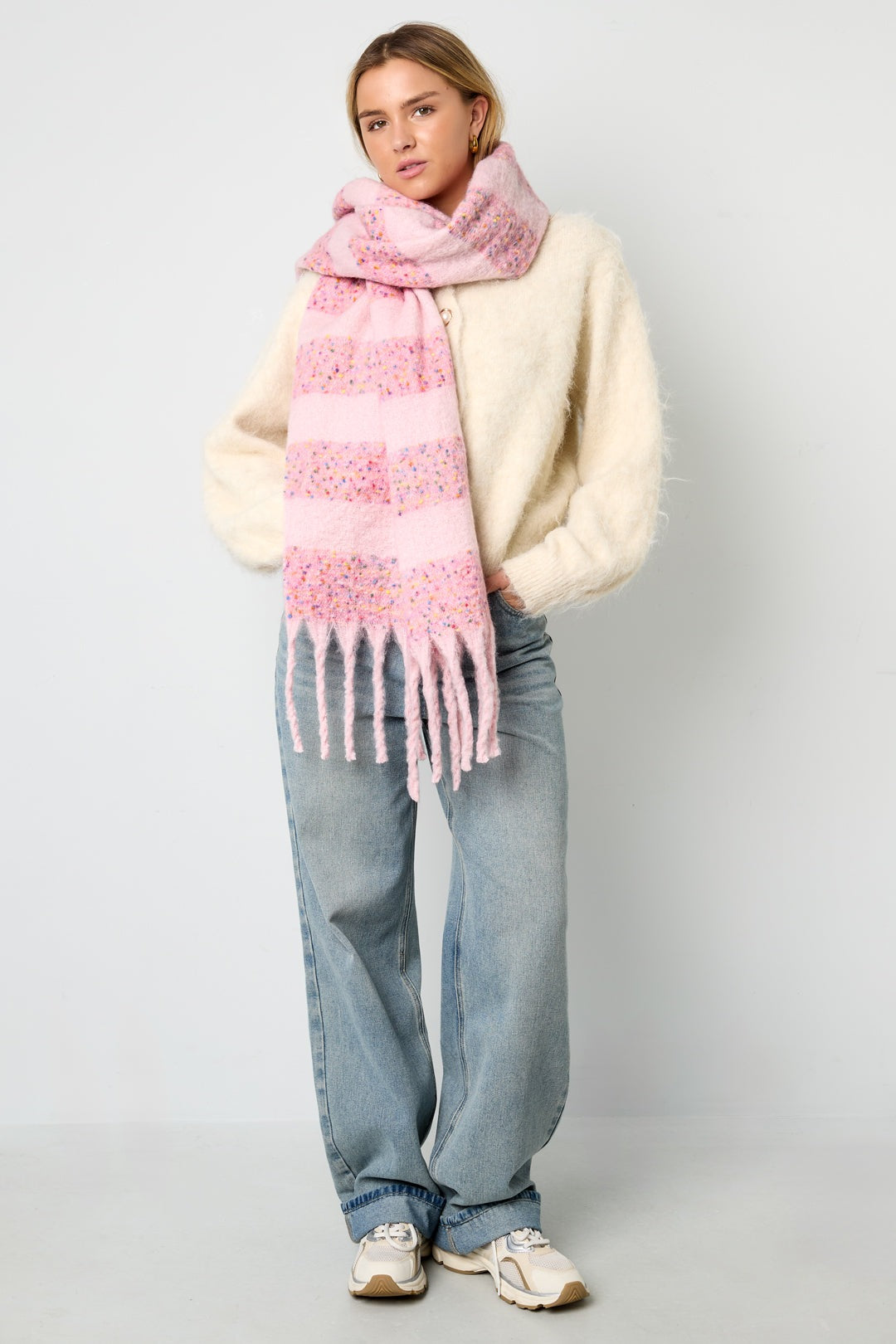 Model wearing a pastel pink scarf with confetti accents wrapped around the neck, paired with a cream fuzzy sweater and light blue jeans