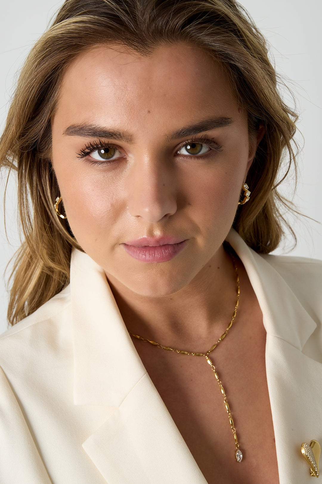 Close-up of front-facing model wearing gold square rhinestone hoop earrings, cream blazer, and gold chain necklace