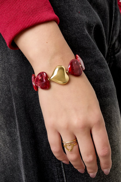 Wrist wearing heart-shaped marbled red acrylic bead bracelet with gold-tone heart accent paired with heart and rhinestone rings