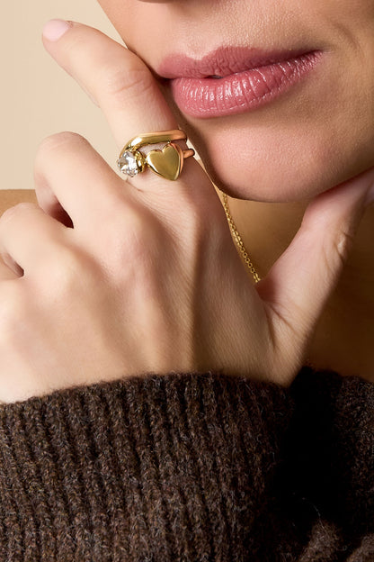 Close-up of woman’s hand showing the gold heart and gemstone ring with soft pink lips and textured brown sweater