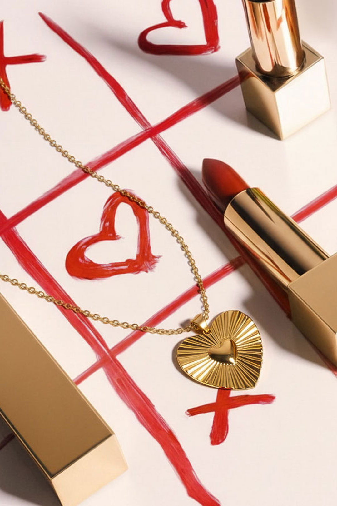 Close-up of gold heart pendant necklace with sunburst pattern placed on tic-tac-toe game grid with lipstick props