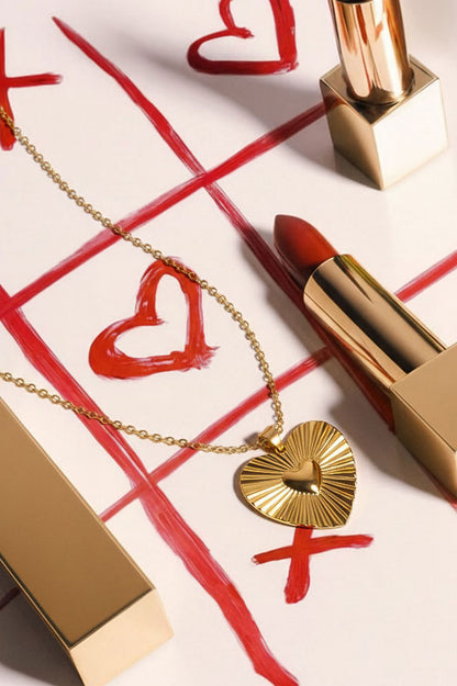 Close-up of gold heart pendant necklace with sunburst pattern placed on tic-tac-toe game grid with lipstick props