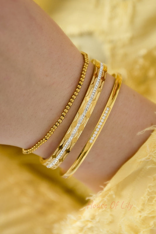 Close-up of a woman's wrist wearing a set of three gold-tone bangles: one beaded, one textured with crystals, and one polished with crystal accents against a yellow fabric background