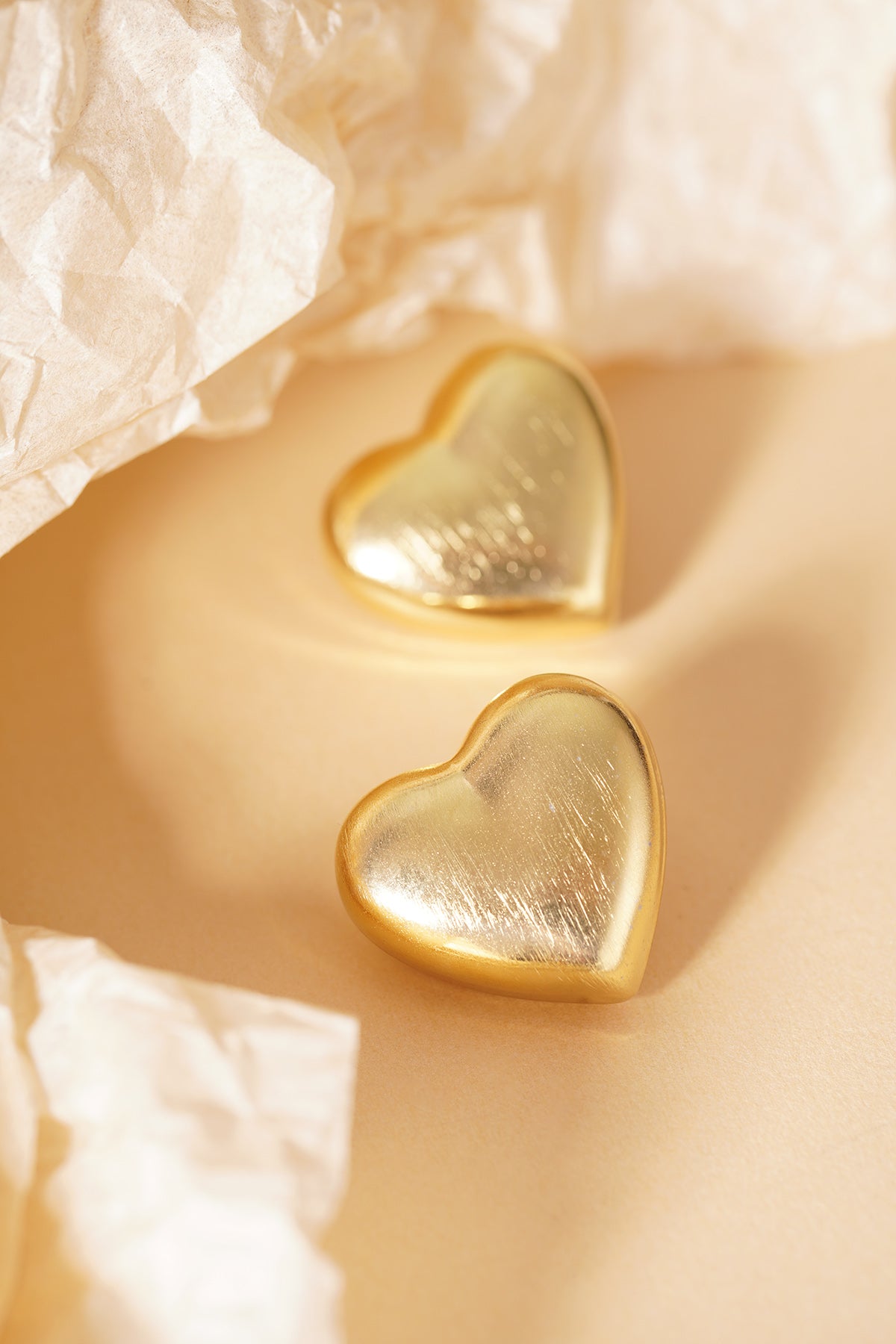Pair of gold matte heart stud earrings on crumpled tissue paper background highlighting soft finish