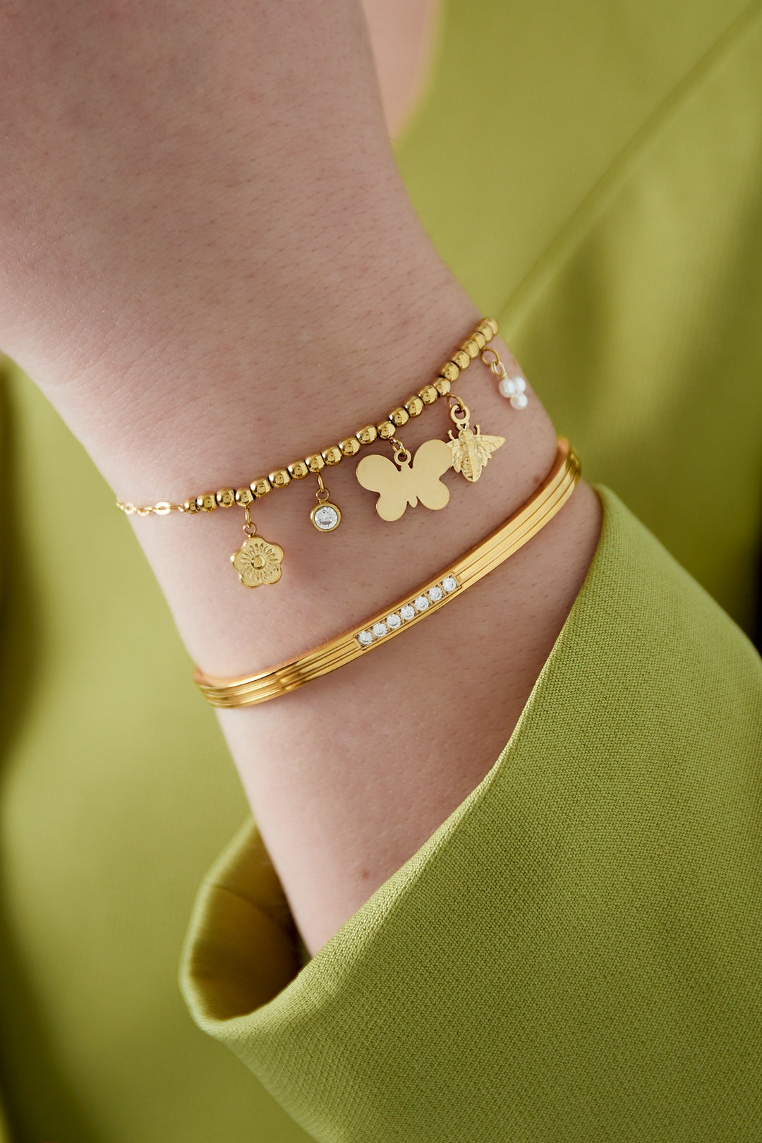 Close-up of gold-tone charm bracelet with textured beads and dangling butterfly, bee, flower, and crystal charms worn on wrist with lime green sleeve