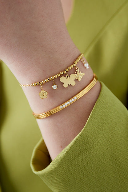 Close-up of gold-tone charm bracelet with textured beads and dangling butterfly, bee, flower, and crystal charms worn on wrist with lime green sleeve