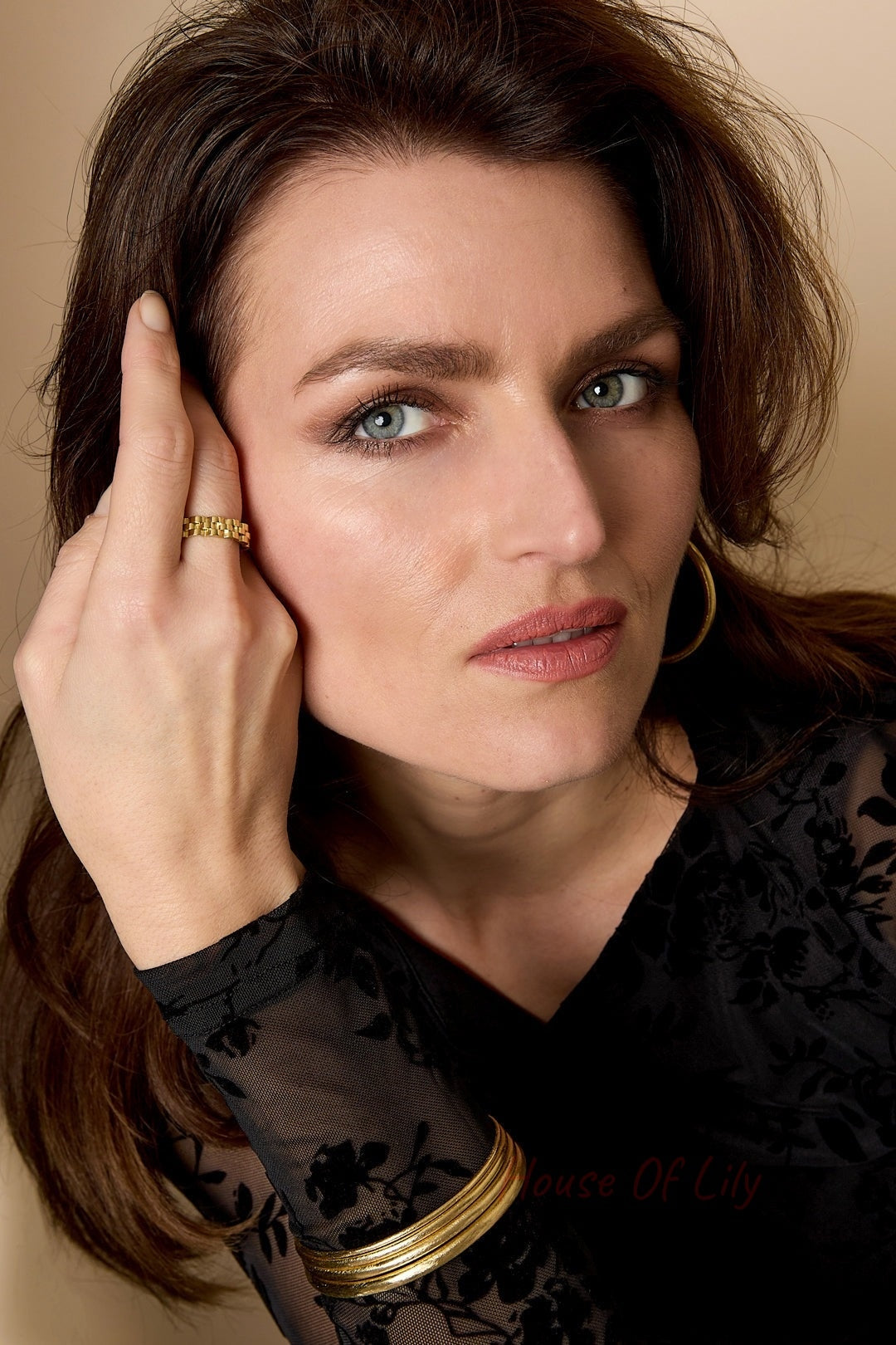 Close-up portrait of woman wearing a gold ring, hoop earrings, and multiple gold bangles on wrist, styled with black floral sheer top
