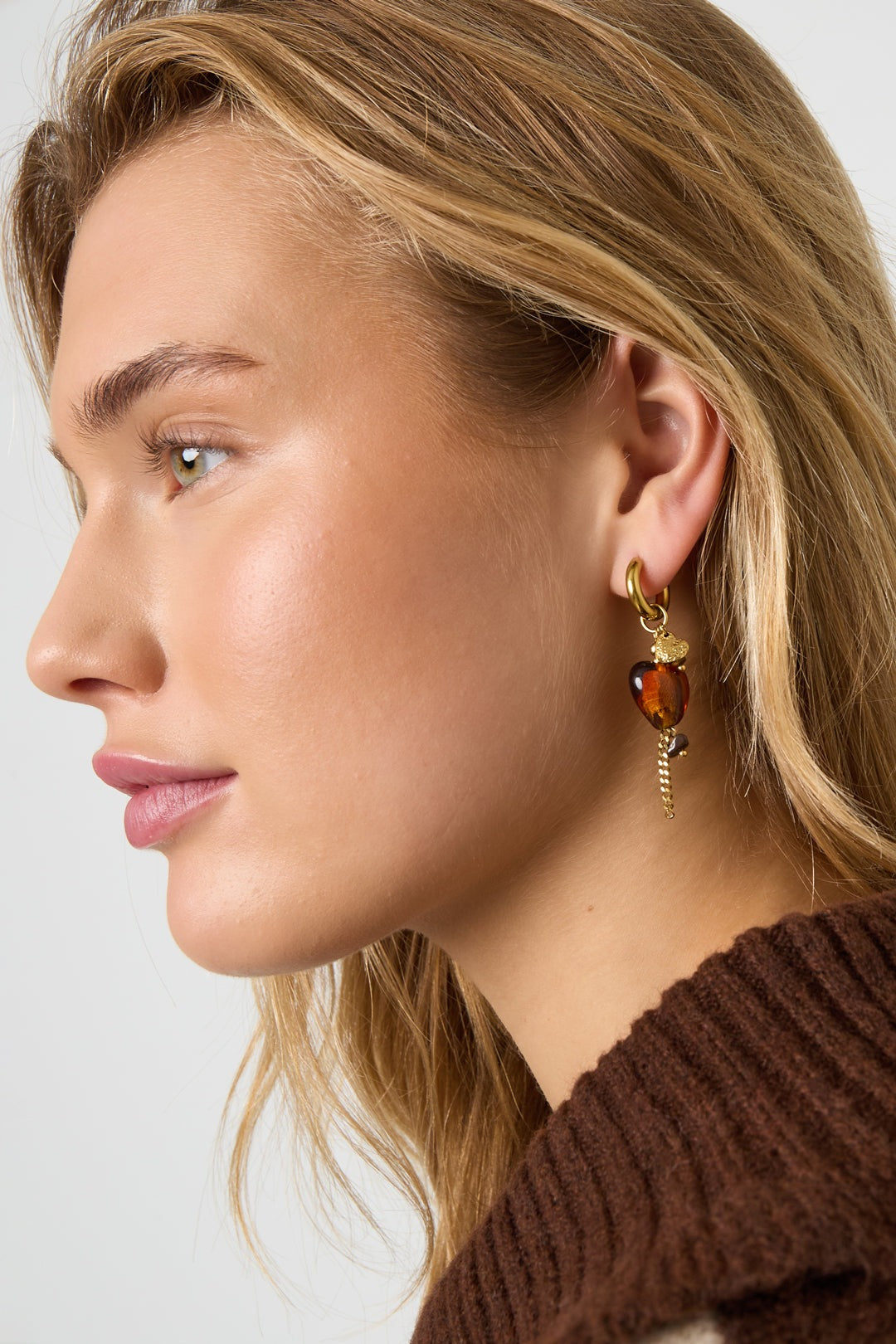 Close-up side profile of a woman wearing gold hoop earring with amber heart charm and chain detail