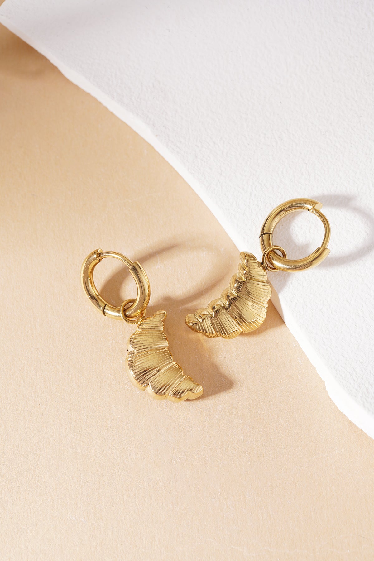 Gold hoop earrings featuring textured, croissant-shaped charms, set against a two-toned tan and white background.