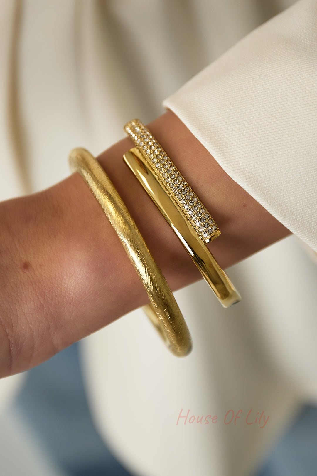 Gold-tone crystal bangle worn stacked with two other gold bracelets on wrist with neutral sleeve