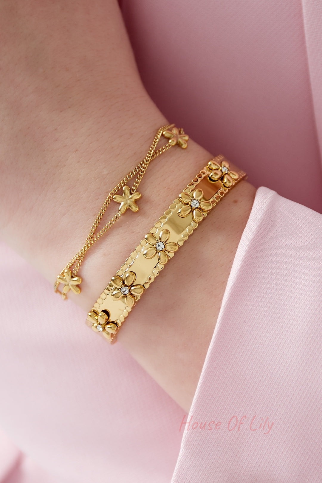 Woman's wrist wearing two gold bracelets, one floral bangle and one chain bracelet with matching flower charms, paired with pink sleeves