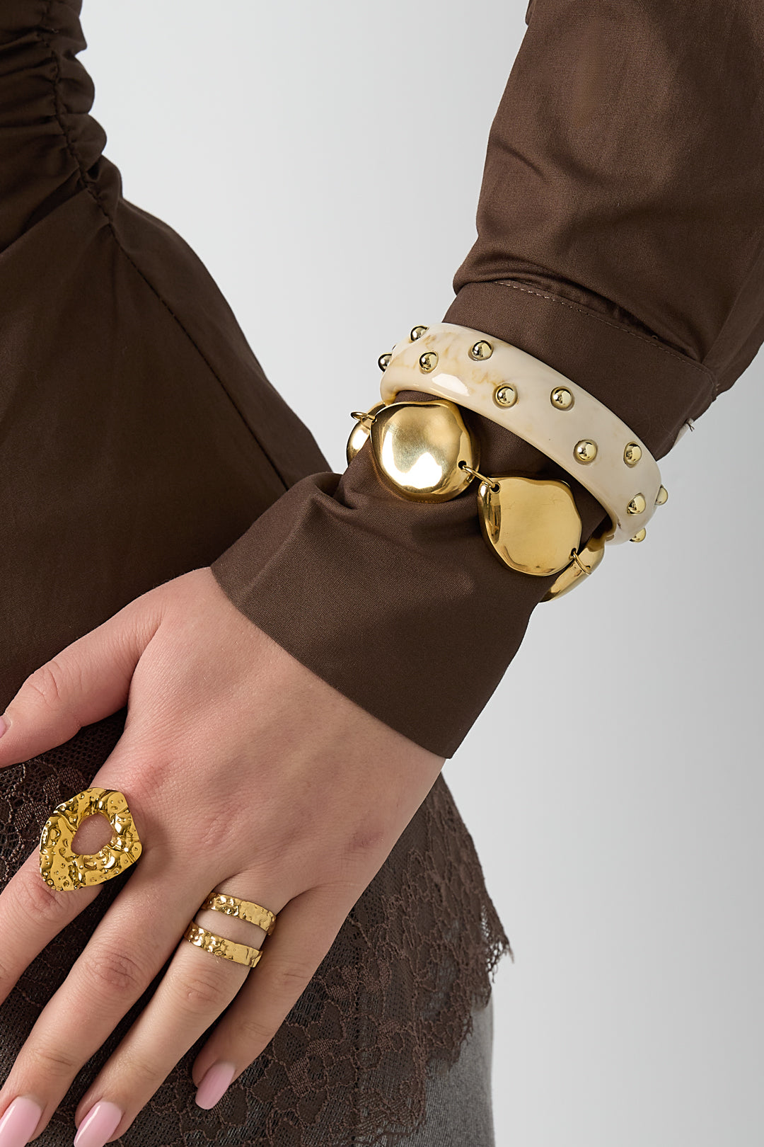 Gold chunky metal bracelet styled on wrist alongside gold textured rings and a brown lace-trim top
