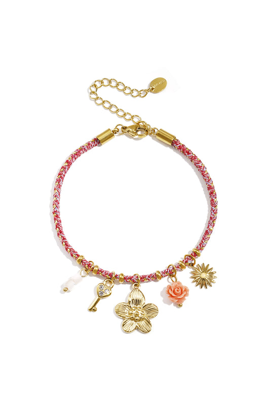 Close-up shot of a charm bracelet with pink woven cord and gold charms including a flower, key, rose, sunburst, and bead on a white background