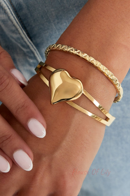 Closeup of two gold bangles on woman's wrist, one with a textured geometric design and one with a heart detail, styled with light denim