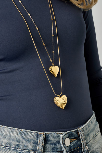 Close-up shot of the layered gold heart pendant necklaces showcasing the textured heart on light-wash denim and navy blue top